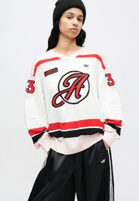Pull de style hockey en blanc avec des accents rouges, noirs et roses. Présente un grand logo, des manches texturées et un design à col en V.