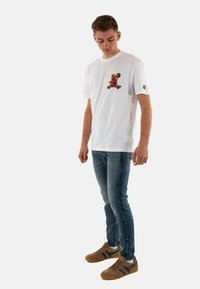 La camiseta blanca de manga corta presenta un gráfico colorido de un oso. Se lleva con unos jeans ajustados de color azul oscuro y zapatillas marrones con acentos azules.