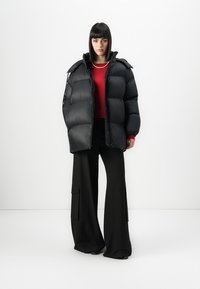 Vrouw in een zwarte pufferjack, rode sweater, zwarte wijde cargo-broek en een korte parelketting, staande tegen een witte achtergrond.