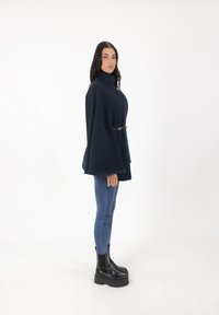 Capa azul marino con un cuello alto y cintura ceñida. Combinada con jeans ajustados azules y botas negras de tobillo de suela gruesa. Textura de tela suave.