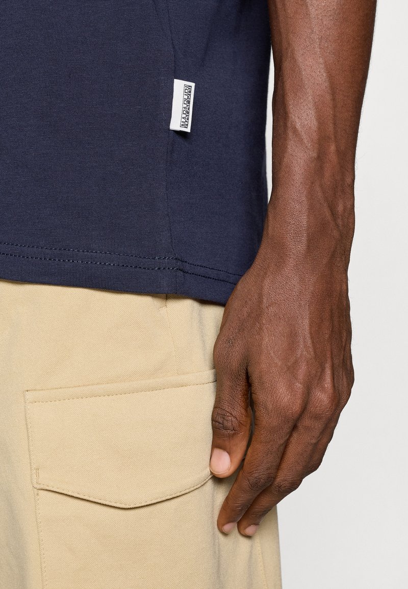 T-shirt bleu marine à manches courtes en coton, avec une étiquette sur le côté et un ourlet droit, associé à un pantalon cargo beige clair avec une poche latérale.