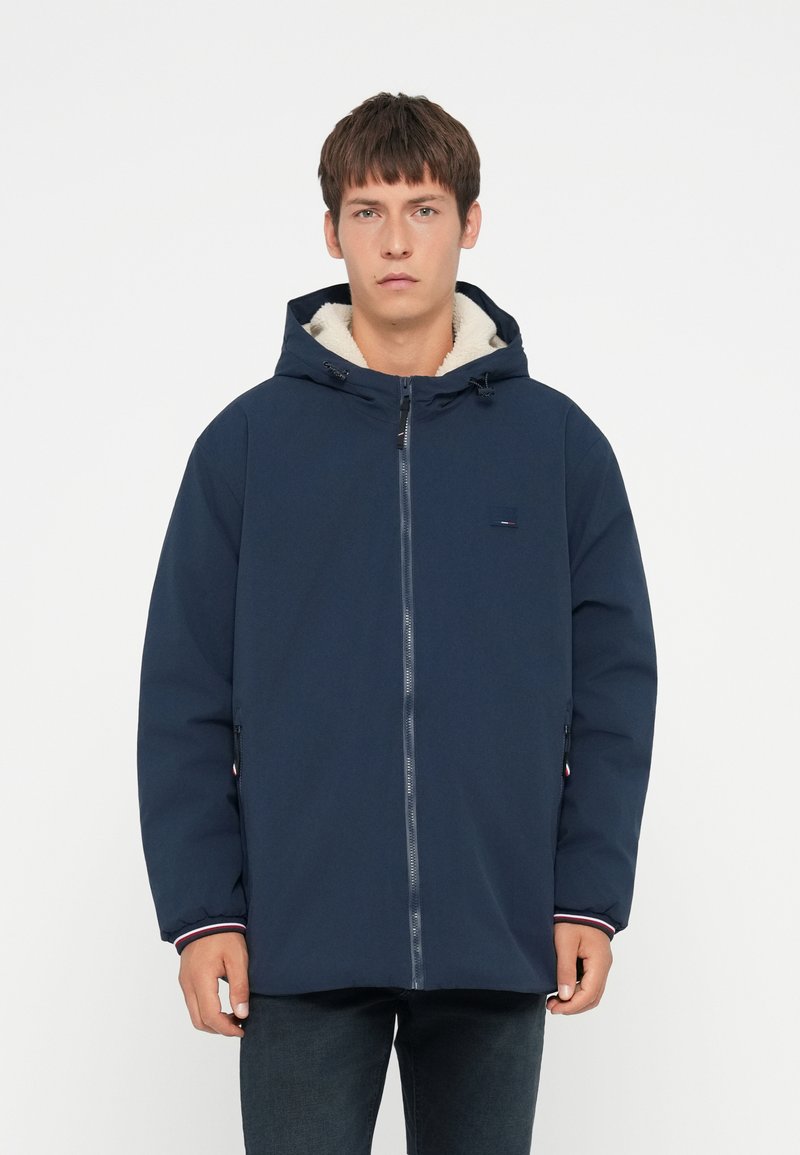 Giacca con cappuccio blu navy, con zip anteriore, foderata in morbido tessuto crema. Presenta polsini elasticizzati e tasche laterali. Dettaglio logo discreto.