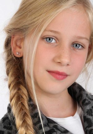 Silberne, herzförmige Ohrringe, geflochtenes blondes Haar, klare blaue Augen, helle Haut mit Sommersprossen, trägt eine graue gemusterte Jacke.
