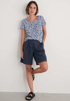 Kortærmet mønstret skjorte i blå, grønne og lilla nuancer med en nøglehulldetalje, parret med marineblå linnedshorts og sorte sandaler.