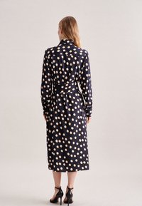 Vestido de lunares en azul marino con grandes lunares crema, mangas largas, cinturón en la cintura y largo a media pantorrilla. Tejido suave con un corte ajustado.