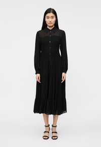 Čierne dlhé šaty s dlhým rukávom, golierom, nažehleným živôtikom, vrstvenou sukňou a volánovým lemom. Hladká látka so zapínaním na gombíky vpredu.