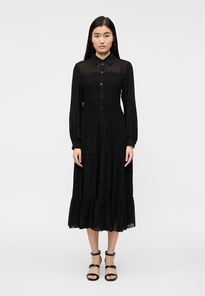 Čierne dlhé šaty s dlhým rukávom, golierom, nažehleným živôtikom, vrstvenou sukňou a volánovým lemom. Hladká látka so zapínaním na gombíky vpredu.
