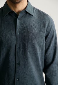 Gestructureerde donkergroene shirt met een knoopsluiting aan de voorkant, spitse kraag en een borstzak aan de linkerkant. Bevat een subtiele gekreukte stofontwerp en ronde knopen.