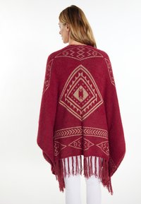 Puncho dengan pola merah dan desain geometris emas, dengan pinggiran hem yang bergerigi dan tekstur lembut. Dipakai di atas celana putih, menunjukkan kontras dan gaya.