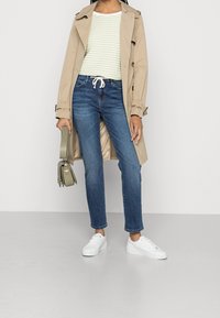 Trench-coat beige sur un haut rayé vert clair, jean bleu et baskets blanches. Tenant un petit sac à main vert olive.