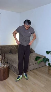 Homme aux cheveux tressés, portant un polo gris, un pantalon noir et des baskets vertes, debout dans un salon en ajustant son pantalon près d'un canapé marron et d'une plante verte.