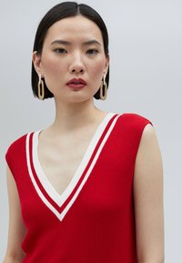 Röd ribbad ärmlös topp med djup V-ringning, försedd med vita randiga detaljer. Modellen bär iögonfallande guld hoopsörhängen. Slät textur.