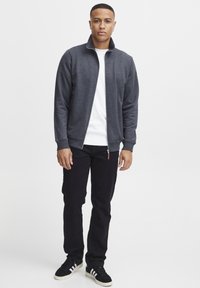 Chaqueta de cremallera gris oscuro con cuello, llevada sobre una camiseta blanca, combinada con pantalones negros y zapatillas negras con rayas blancas.