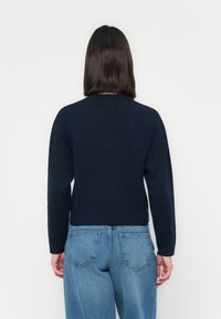Granatowy sweter z dzianiny o teksturowanym wzorze, okrągłym dekolcie i długich rękawach, zestawiony z jasnoniebieskimi dżinsami.