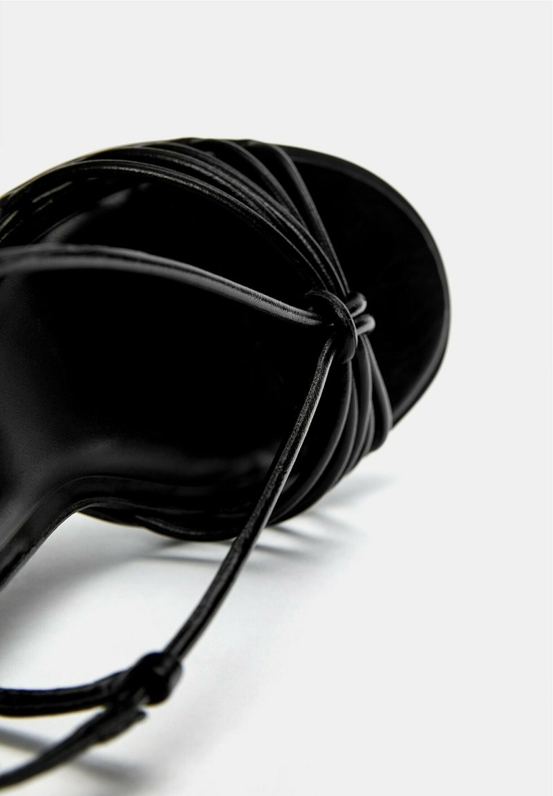 Sandal de tacón alto negro con múltiples correas delgadas, textura de cuero suave y diseño de punta cerrada. Suela de goma visible por debajo.