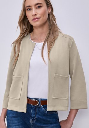 Femme portant une veste beige à ouverture frontale avec de grandes poches avant, un haut blanc, une ceinture marron et un jean bleu sur fond uni.