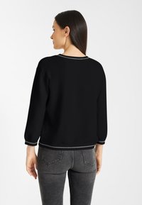 Vrouw met lang, donker haar in een zwarte trui en grijze jeans, staande in profil met haar rug gedeeltelijk gekeerd.