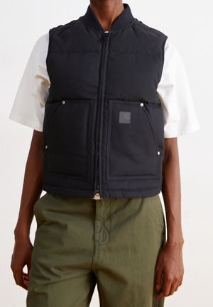 Persoon draagt een zwart gewatteerd vest met ritssluiting en voorzakken over een wit T-shirt met korte mouwen en olijfgroene broek, staand voor een effen achtergrond.