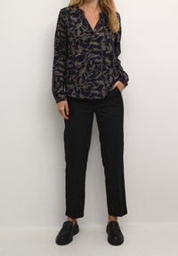 Blouse bleu marine avec motif abstrait beige, col en V, manches longues. Pantalon noir ajusté. Mocassins plats noirs.