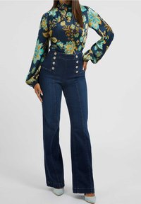 Kvinde iført højtaljede mørkeblå jeans med guldknapper og en langærmet bluse med blomster mønster i blå og guld nuancer.