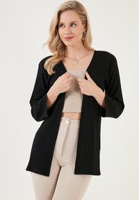 Cardigan léger noir avec un tissu texturé, à l'ouverture devant et aux manches trois-quarts, superposé sur un top court beige et un pantalon beige.