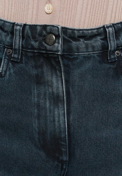 Jeans din denim de culoare albastră închisă, cu nasturi și bucle pentru curea, și o parte a unei cămăși bej cu dungi, cu un singur nasture bej vizibil deasupra.