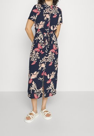 Frau trägt ein marineblaues, kurzärmliges Midi-Kleid mit Knöpfen und rosa Schmetterlings- und Blattmustern, kombiniert mit weißen Sandalen mit offenen Zehen.