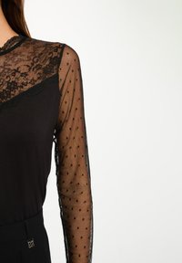Top negro de manga larga con un yugo de encaje con un patrón floral y mangas transparentes de lunares. Tela suave con una silueta ajustada.