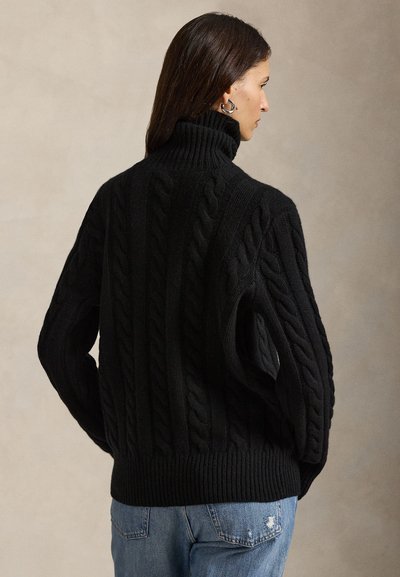 Femme aux longs cheveux foncés portant des boucles d’oreilles argentées en forme d’anneaux, un pull épais en maille torsadée noire à col roulé, et un jean bleu avec une petite déchirure.