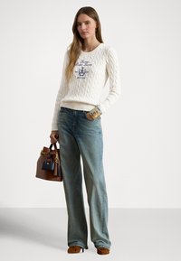 Krem pulover z vzorcem iz pletenja z modrim izvezenim logotipom, v kombinaciji s svetlo modrimi kavbojkami širokih hlačnic. Rjava torbica s cofki in zlatimi poudarki, rjavi čevlji.