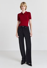 Donna in piedi che indossa una polo rossa aderente a maniche corte, jeans neri a vita alta e gamba larga con un ginocchio strappato e scarpe nere con punta.