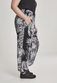 Personne portant un pantalon large à motifs noir et blanc avec taille élastique et des baskets noires, main dans la poche, debout de côté.
