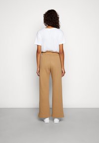 Brązowe spodnie szerokie z elastycznym pasem, w połączeniu z gładką białą koszulką. Model tyłem, w białych sneakersach.