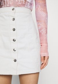 Valkoinen denim-hame, jossa on etunappikiinnitys, ja se on varustettu hopeanvärisillä yksityiskohdilla sekä sileällä pinnalla. Vyötärö on hieman korkeavyötäröinen, ja siinä on hienovaraisia saumoja.