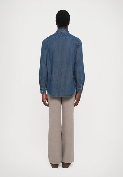 Chemise en denim avec un col button-down, des manches longues et un ourlet arrondi, portée sur un pantalon évasé gris clair.