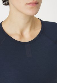 Marineblaue Bluse mit rundem Ausschnitt, ausgestattet mit einem strukturierten Etikett mit "Rapha". Weicher Stoff mit sichtbaren Nähten.