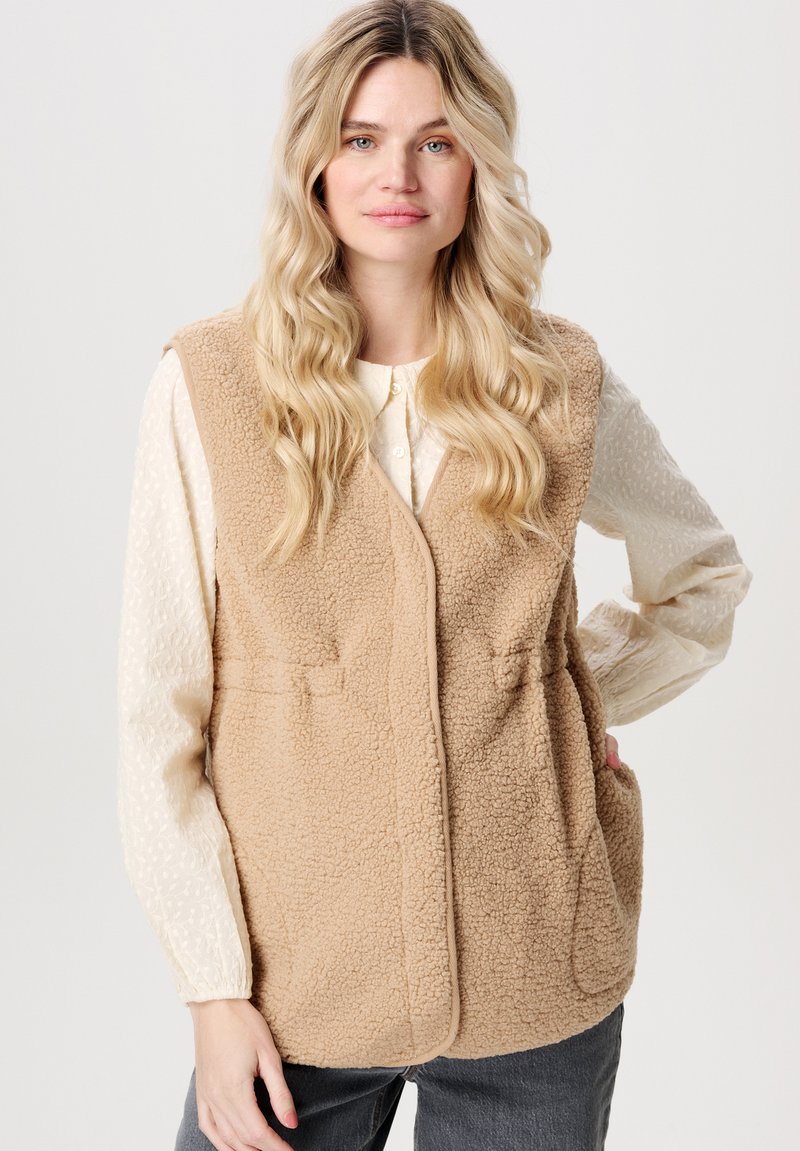 Beige konstfuskpälsväst med en avslappnad passform, öppen front och två framfickor. Bärs över en texturerad krämfärgad blus, ihop med mörka jeans.