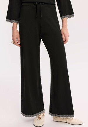 Kobieta nosząca czarne spodnie o szerokich nogawkach z białym paskowanym wykończeniem na mankietach oraz kremowe sznurowane buty, stojąca na jednolitym tle.