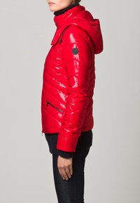 Röd vadderad jacka med blank yta, med avtagbar huva, dragkedjefickor och en svart loggopatch på ärmen.