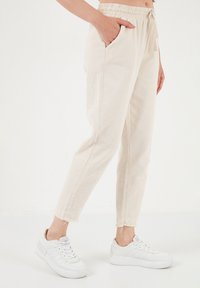 Beige lightweight jogger pants with an elastic waistband and drawstring. Features side pockets and frayed hems, worn with white sneakers.
