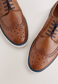 Braune Lederschuhe mit perforierten Brogue-Details, dunkelblauen Schnürsenkeln und einer weißen Sohle mit blauem Akzent am Rand.