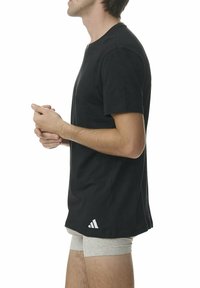 T-shirt noir à manches courtes en coton, avec un col rond et un accent logo blanc sur l'ourlet inférieur. Porté par-dessus un sous-vêtement gris clair.