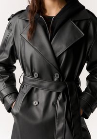 Manteau croisé en similicuir noir avec larges revers, taille ceinturée, poches latérales et accents de boutons, présentant une texture lisse.