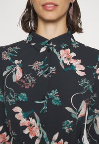 Blusa a fiori in tessuto scuro, con chiusura a bottoni sul davanti, colletto e una varietà di design botanici in rosa e verde.