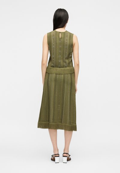 Γυναίκα με μακριά σκούρα μαλλιά, ντυμένη με ένα αμάνικο πλεκτό φόρεμα σε ελαιί πράσινο με κρόσια, στέκεται με την πλάτη, μαύρα σανδάλια με λευκά τακούνια block.