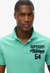 Polo léger en tissu texturé de couleur bleu-vert. Doté d'une patte de boutonnage à trois boutons, avec un logo brodé bleu marine "SUPERDRY Athletic 54" sur la poitrine.