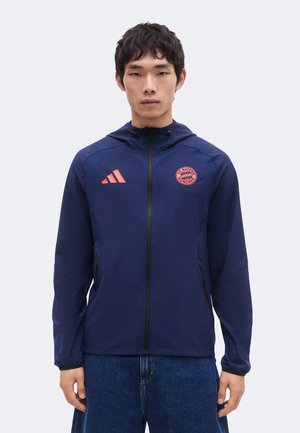 Mann trägt eine marineblaue Kapuzenjacke mit Reißverschluss, pinkem Adidas-Logo und FC Bayern München Wappen, kombiniert mit blauen Jeans.