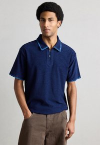 Μπλε ναυτικό πόλο πουκάμισο με υφασμάτινη υφή, αντίθετο μπλε γιακά και περιθώριο στα μανίκια, τριπλό κουμπί στη λαιμόκοψη, κοντά μανίκια και χαλαρή εφαρμογή.