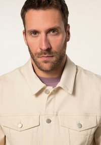 Chaqueta vaquera beige con botones plateados, collar clásico y dos bolsillos en el pecho. Llevada sobre una camiseta de color púrpura claro.
