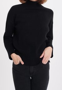 Czarny sweter z golfem o luźnym kroju, z prążkowanymi mankietami i dołem. Materiał wydaje się miękki i teksturowany. Noszony z czarnymi jeansami.